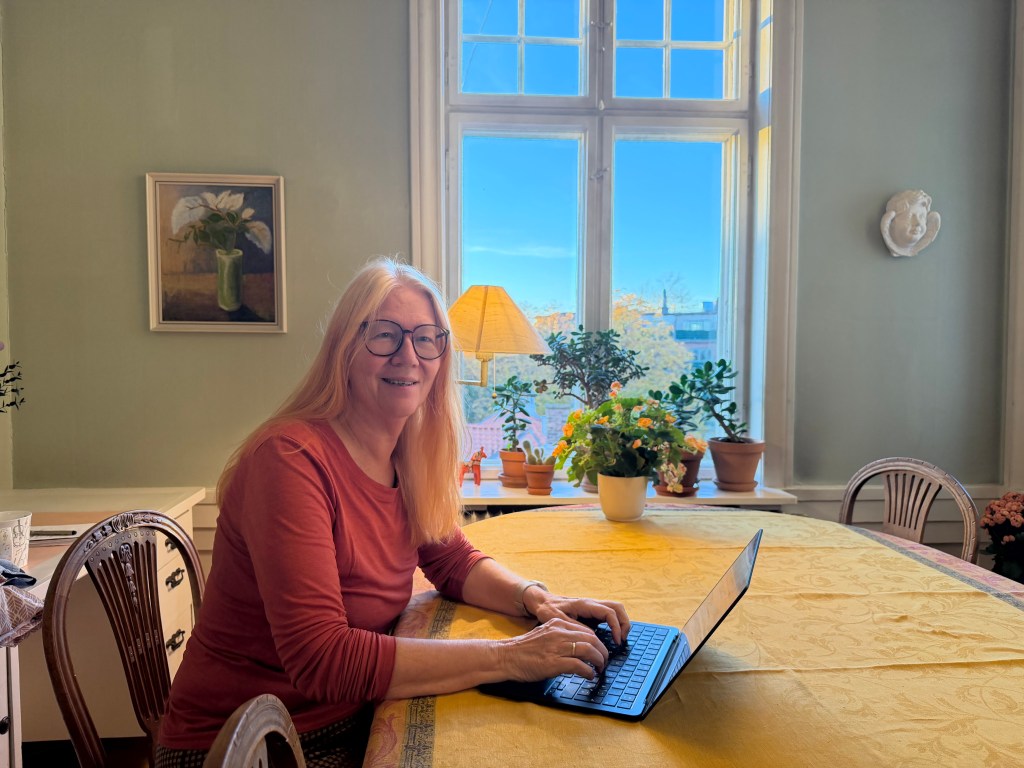 Jannike sittende med PC ved spisestuebordet. Blå himmel synlig gjennom vinduet, og planter i vindusposten.